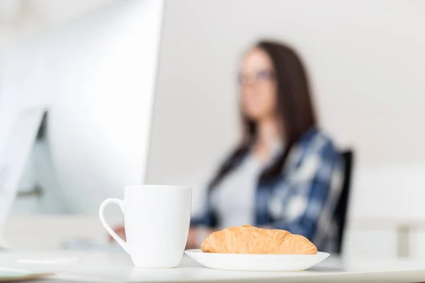 Uzavřete šálek kávy nebo čaje a čerstvý čokoládový croissant vedle mladé ženy pracující na stolním počítači. — Stock fotografie