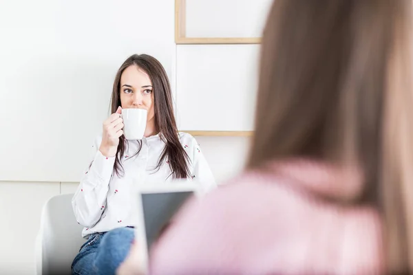 Dvě mladé moderní samice popíjí kávu a používají tabletové zařízení v moderní kanceláři. — Stock fotografie