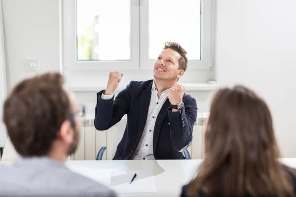 Χαρούμενος επιχειρηματίας με τη νίκη χειρονομία σε μια συνάντηση με τους συναδέλφους του που κάθονται στο γραφείο στο γραφείο. — Φωτογραφία Αρχείου