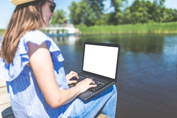 Krásná hipsterová holka sedí na doku a pracuje na laptopu. Jezero nebo řeka v pozadí. Koncepce práce a cestování. — Stock fotografie