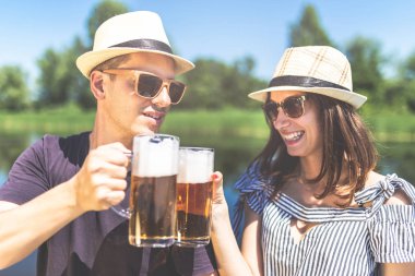 Arka planda güzel göle karşı bira bardakları olan neşeli genç bir hippi çift..