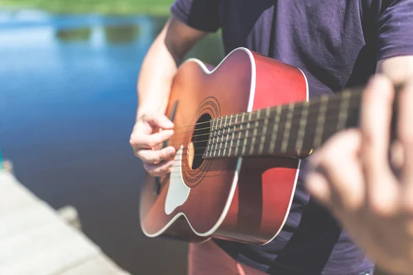 Close up of man playing acoustic guitar against beautiful lake or river. — Stock Photo, Image
