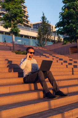 Modern freelancer akıllı telefon konuşuyor ve gün batımında iş binası önünde adımlar üzerinde otururken dizüstü bilgisayar kullanarak.