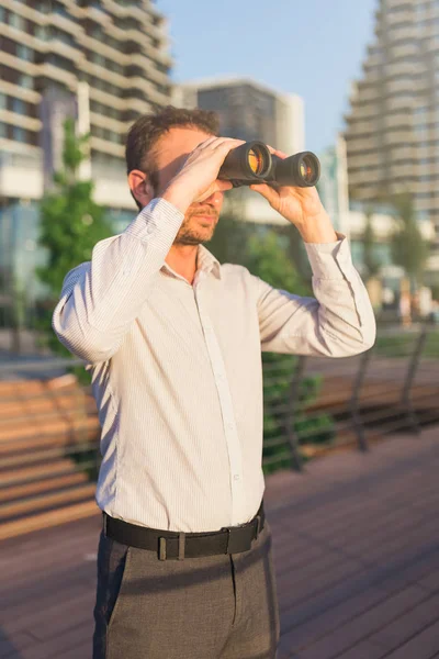 Business person looking through binoculars in front of business buildings.