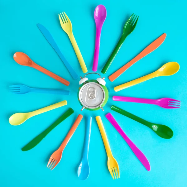 Top view of multicolored plastic spoons, forks and knives with aluminum can in form of alarm clock isolated on blue. Food and drink time abstract.