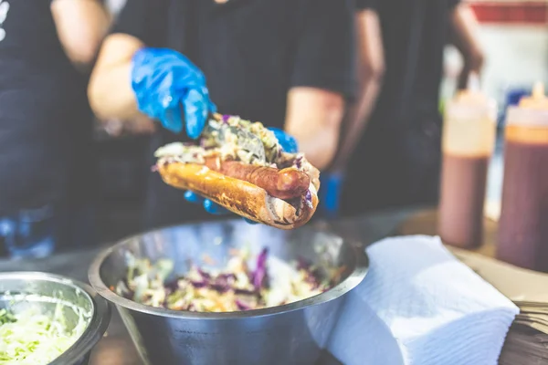 Gros plan des mains du chef ajoutant des légumes sur un hot-dog frais avec des saucisses grillées . — Photo
