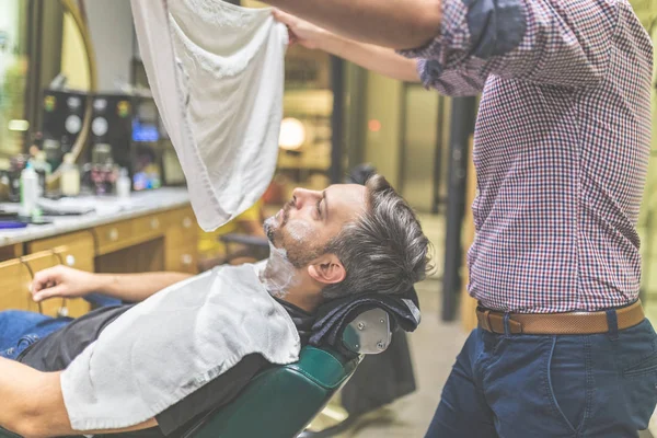Парикмахер готовит клиента к бритью полотенцем. Человек, посещающий парикмахерскую . — стоковое фото