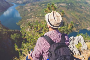 Havalı bir gezgin kayalıklarda oturuyor ve tepelerle çevrili göle ya da nehre bakıyor..