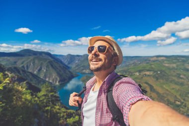 Cheerful man traveler with straw hat and sunglasses taking selfie against beautiful valley. Travel and nature explore concept. Space for copy. clipart