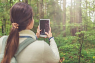 Boş ekran tableti kullanan sırt çantalı bir kadın yürüyüşçü. Arka planda doğa manzarası.