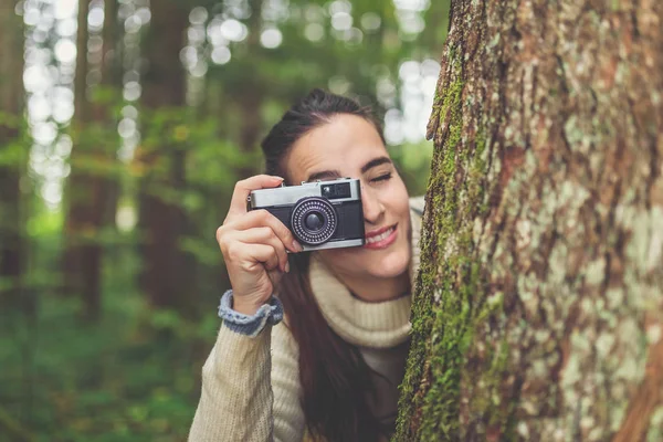 Portrait of beautiful woman using retro film camera behind tree in the woods. — ストック写真