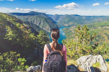 Dağın tepesindeki kayalarda oturan genç bayan gezgin vadi gölü ya da tepeli nehrin manzarasının tadını çıkarıyor..