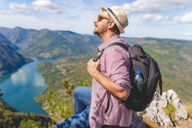 Havalı bir gezgin kayalıklarda oturuyor ve tepelerle çevrili göle ya da nehre bakıyor..