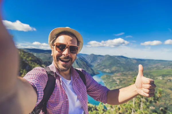 Turista de moda tomando fotos de sí mismo mientras está de pie en la colina sobre el cañón y las montañas y mostrando los pulgares hacia arriba . —  Fotos de Stock