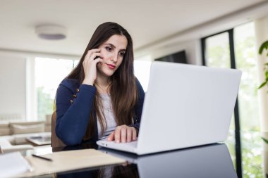 Evde çalışan genç bir kadın, cep telefonu ve dizüstü bilgisayar kullanıyor. Çevrimiçi toplantı kavramı.