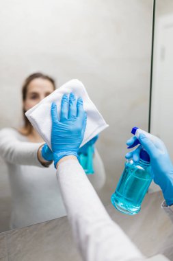 Genç ve güzel bir bayan koruyucu yüz maskesi takıyor ve banyoda ayna temizliyor. Coronavirus koruma.