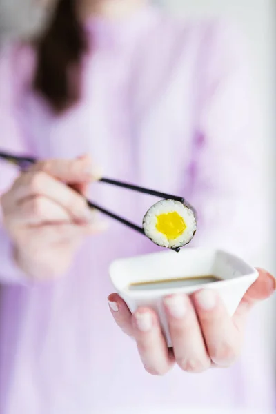 Female dipping tasty sushi maki or uramaki with chopsticks into bowl with soy sauce.