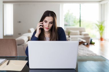İş kadını akıllı telefondan konuşuyor ve evde dizüstü bilgisayar kullanıyor. Uzak çalışma konsepti.