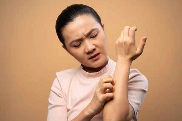 Asian woman was sick with irritate itching her skin, scratching her skin, standing isolated on beige background.