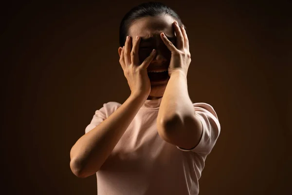 Asian woman scared screaming covering face by her hands standing isolated on beige background. Low key.
