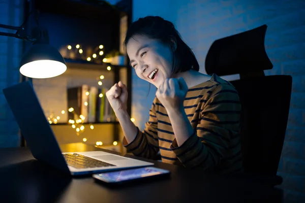 Asian woman happy smiling working on a laptop at the night at home and making winning gesture. WFH. Work from home avoid COVID 19 concept.