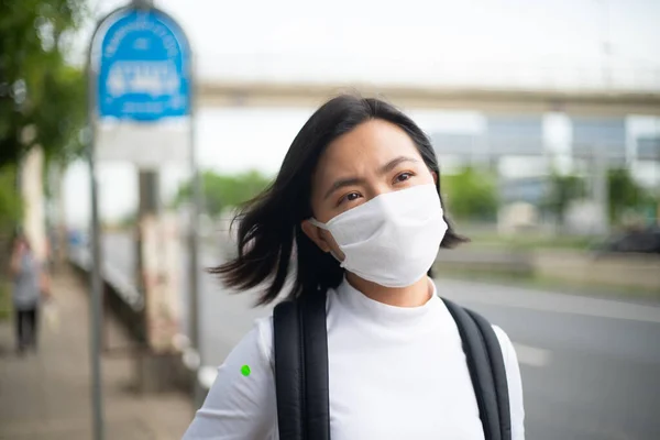 Asian woman wearing protective face mask prevention virus and pollution, standing at bus stop. New lifestyle with Corona Virus COVID-19.