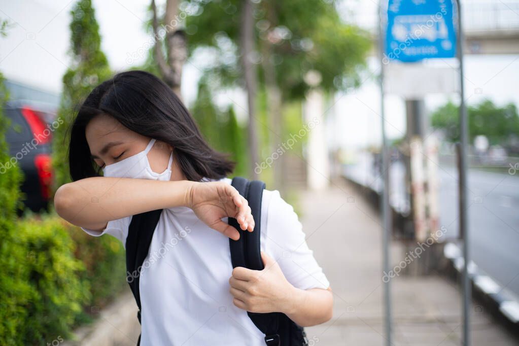 Asian woman wearing protective face mask prevention virus and pollution, coughing and standing at bus stop. New lifestyle with Corona Virus COVID-19.