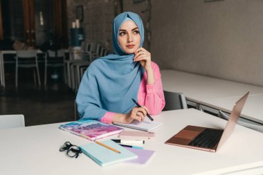 Genç, modern, tesettürlü Müslüman bir kadın ofis odasında dizüstü bilgisayarla çalışıyor. İnternette eğitim görüyor, uzaktan çalışıyor.