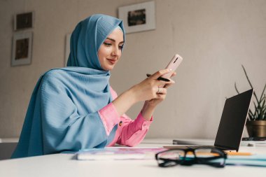 Genç, modern, tesettürlü Müslüman bir kadın ofis odasında dizüstü bilgisayarla çalışıyor. İnternette eğitim görüyor, uzaktan kumandalı çalışıyor. Smatphone kullanıyor.