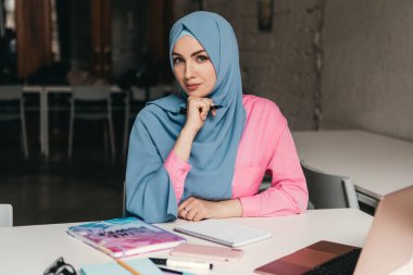 Genç, modern, tesettürlü Müslüman bir kadın ofis odasında dizüstü bilgisayarla çalışıyor. İnternette eğitim görüyor, uzaktan çalışıyor.