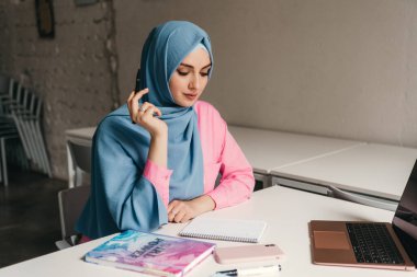 Genç, modern, tesettürlü Müslüman bir kadın ofis odasında dizüstü bilgisayarla çalışıyor. İnternette eğitim görüyor, uzaktan çalışıyor.