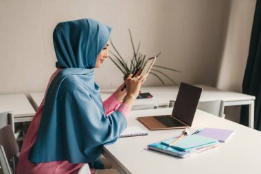 Genç, modern, tesettürlü Müslüman bir kadın ofis odasında dizüstü bilgisayarla çalışıyor. İnternette eğitim görüyor, uzaktan kumandalı çalışıyor. Smatphone kullanıyor.