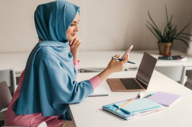 Genç, modern, tesettürlü Müslüman bir kadın ofis odasında dizüstü bilgisayarla çalışıyor. İnternette eğitim görüyor, uzaktan kumandalı çalışıyor. Smatphone kullanıyor.