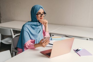 Genç, modern, tesettürlü Müslüman bir kadın ofis odasında dizüstü bilgisayarla çalışıyor. İnternette eğitim görüyor, uzaktan kumandalı çalışıyor. Smatphone kullanıyor.