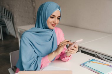 Genç, modern, tesettürlü Müslüman bir kadın ofis odasında dizüstü bilgisayarla çalışıyor. İnternette eğitim görüyor, uzaktan kumandalı çalışıyor. Smatphone kullanıyor.