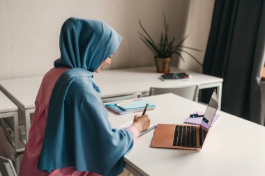 Genç, modern, tesettürlü Müslüman bir kadın ofis odasında dizüstü bilgisayarla çalışıyor. İnternette eğitim görüyor, uzaktan çalışıyor.