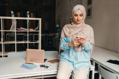 Genç, modern, tesettürlü, ofis odasında çalışan, internetten eğitim alan, uzaktan kumandalı, akıllı telefon kullanan Müslüman bir kadın.
