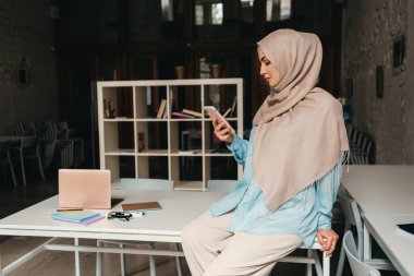 Genç, modern, tesettürlü, ofis odasında çalışan, internetten eğitim alan, uzaktan kumandalı, akıllı telefon kullanan Müslüman bir kadın.