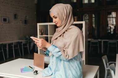 Genç, modern, tesettürlü, ofis odasında çalışan, internetten eğitim alan, uzaktan kumandalı, akıllı telefon kullanan Müslüman bir kadın.