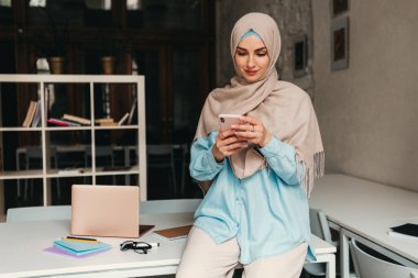 Genç, modern, tesettürlü, ofis odasında çalışan, internetten eğitim alan, uzaktan kumandalı, akıllı telefon kullanan Müslüman bir kadın.