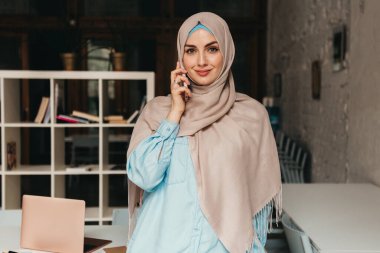 Genç, modern, tesettürlü Müslüman bir kadın ofis odasında çalışıyor, internetten eğitim görüyor, uzaktan kumandalı çalışıyor, akıllı telefondan konuşuyor.