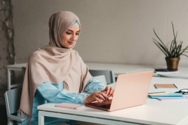 Genç, modern, tesettürlü Müslüman bir kadın ofis odasında dizüstü bilgisayarla çalışıyor, internetten eğitim görüyor, uzaktan kumandalı çalışıyor, daktilo düşünüyor.