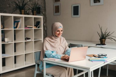 Genç, modern, tesettürlü Müslüman bir kadın ofis odasında dizüstü bilgisayarla çalışıyor, internetten eğitim görüyor, uzaktan kumandalı çalışıyor, daktilo düşünüyor.