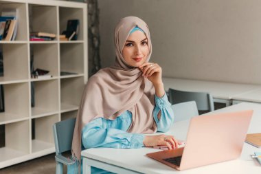 Genç, modern, tesettürlü Müslüman bir kadın ofis odasında dizüstü bilgisayarla çalışıyor. İnternette eğitim görüyor, uzaktan çalışıyor.