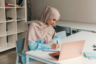 Genç, modern, tesettürlü Müslüman bir kadın ofis odasında dizüstü bilgisayarla çalışıyor. İnternette eğitim görüyor, uzaktan çalışıyor.