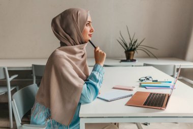 Genç, modern, tesettürlü Müslüman bir kadın ofis odasında dizüstü bilgisayarla çalışıyor. İnternette eğitim görüyor, uzaktan çalışıyor.