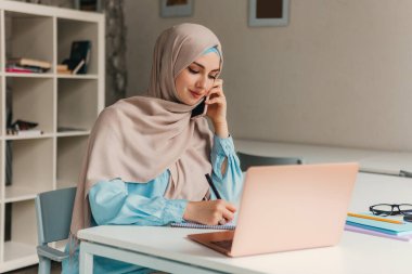Genç, modern, tesettürlü Müslüman bir kadın ofis odasında dizüstü bilgisayarla çalışıyor, internetten eğitim görüyor, uzaktan kumandalı çalışıyor, akıllı telefondan konuşuyor, notlar alıyor.