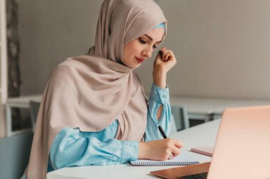 Genç, modern, tesettürlü Müslüman bir kadın ofis odasında dizüstü bilgisayarla çalışıyor. İnternette eğitim görüyor, uzaktan çalışıyor.
