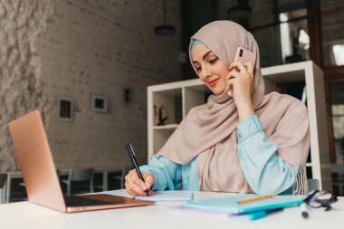 Genç, modern, tesettürlü Müslüman bir kadın ofis odasında dizüstü bilgisayarla çalışıyor, internetten eğitim görüyor, uzaktan kumandalı çalışıyor, akıllı telefondan konuşuyor, notlar alıyor.