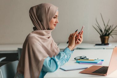 Genç, modern, tesettürlü Müslüman bir kadın ofis odasında dizüstü bilgisayarla çalışıyor, internetten eğitim görüyor, uzaktan kumandalı, akıllı telefon kullanıyor.